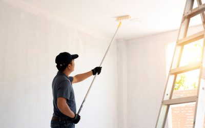 Asian rear view of a male painter drawing a wall with paint roller and a separate tank from a large empty space with wooden stairs.